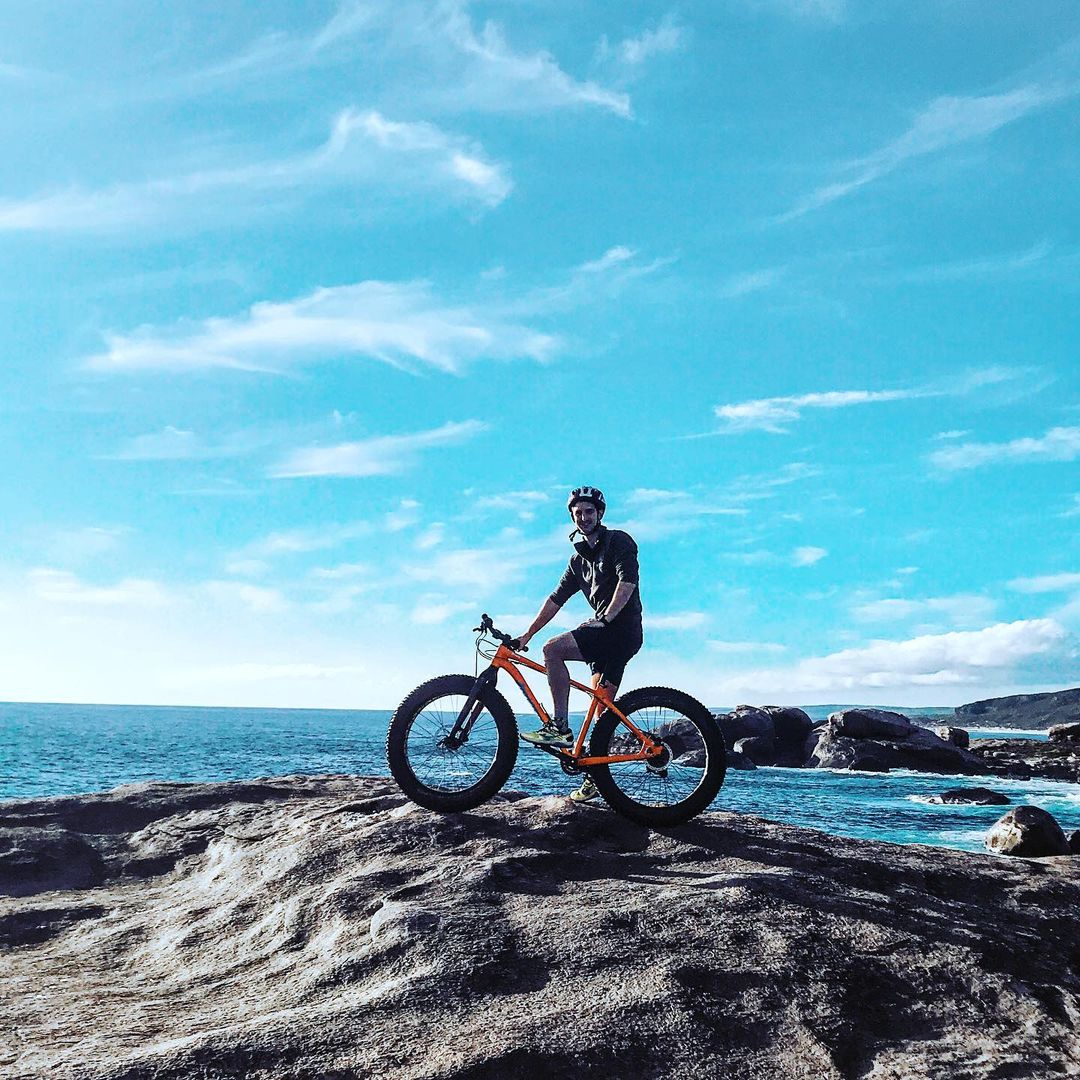 Happy man sitting on fat bike on a granite rock with blue ocean behind.